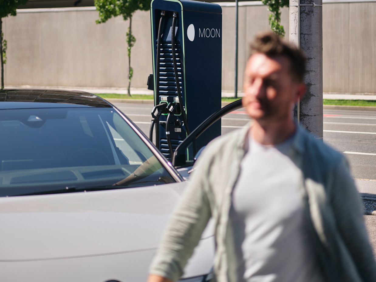 Homem anda na frente de um carro em frente a uma estação de carregamento