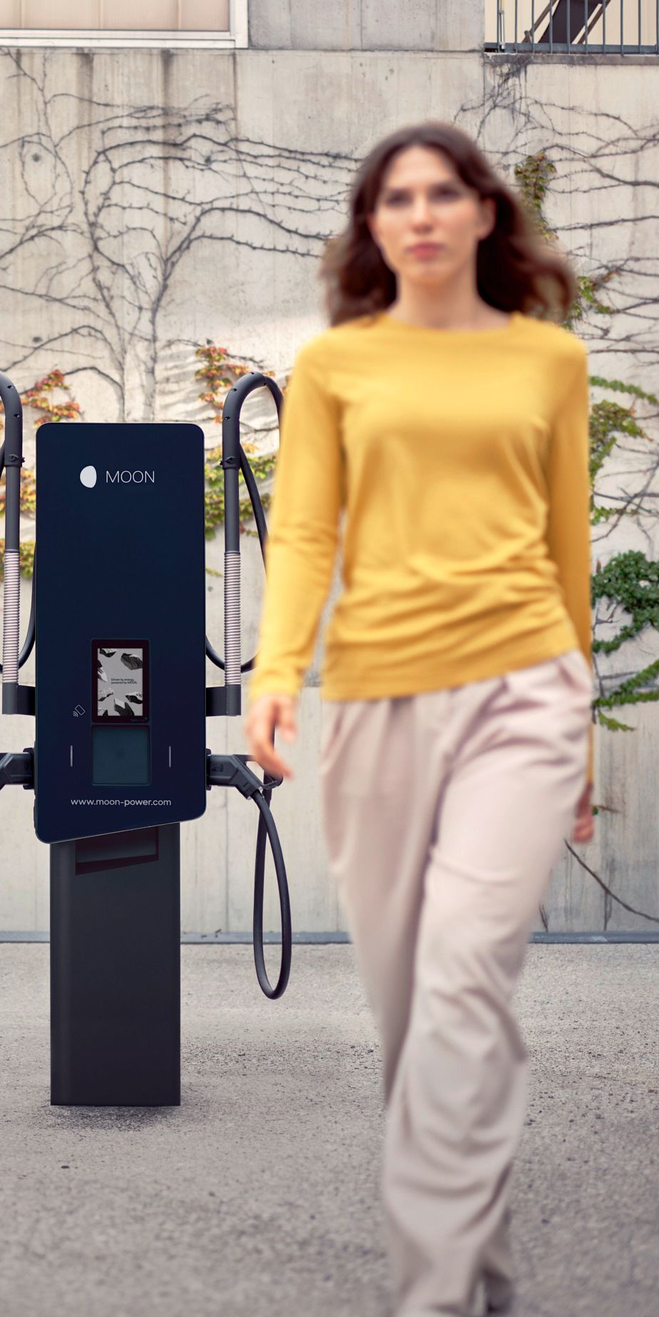 Frau geht vor einer Ladestation vorbei mit einem Auto im Hintergrund