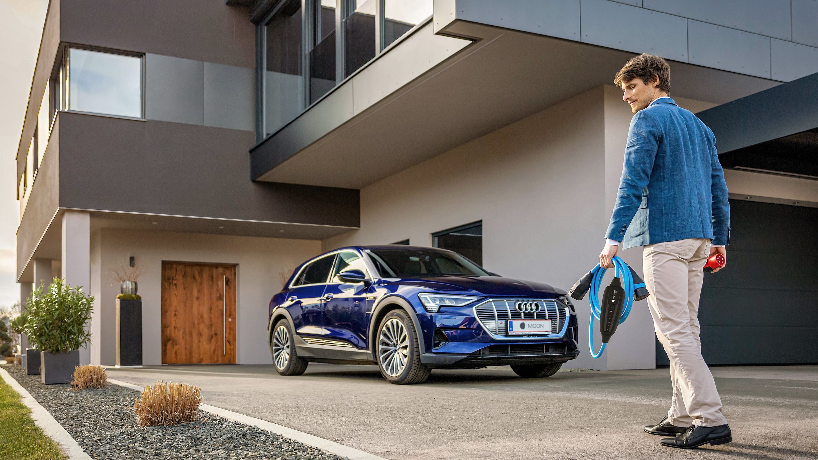 Man walks towards a car near a house with a cable in his hand